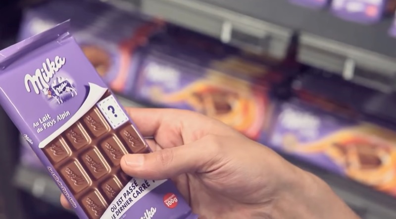 A Milka chocolate bar package with a question mark printed over a missing piece of chocolate. 