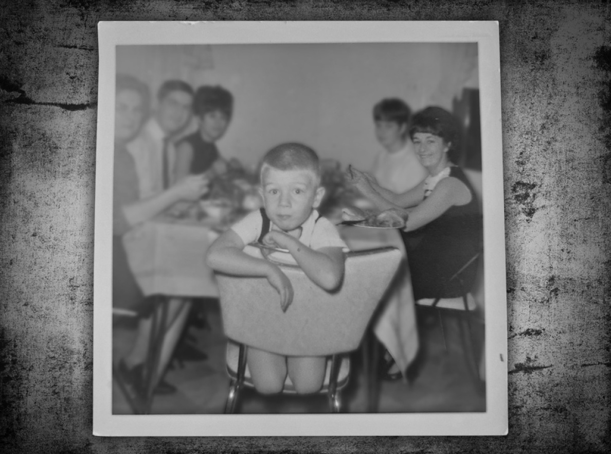 A black and white photograph of Normand Miron as a young boy at the dinner table with his mom (right) and family. 