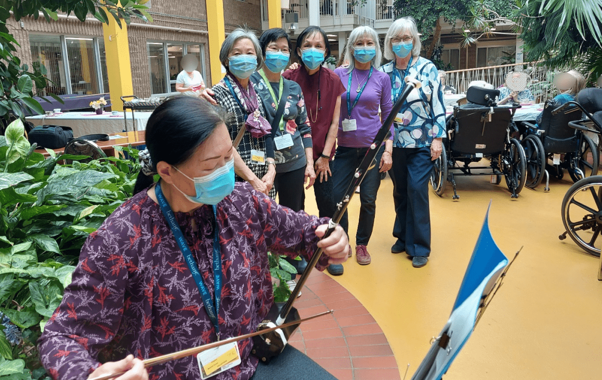 The founder of MPC Foundation poses with participants at a retirement home. A woman in the foreground plays a Chinese violin. 