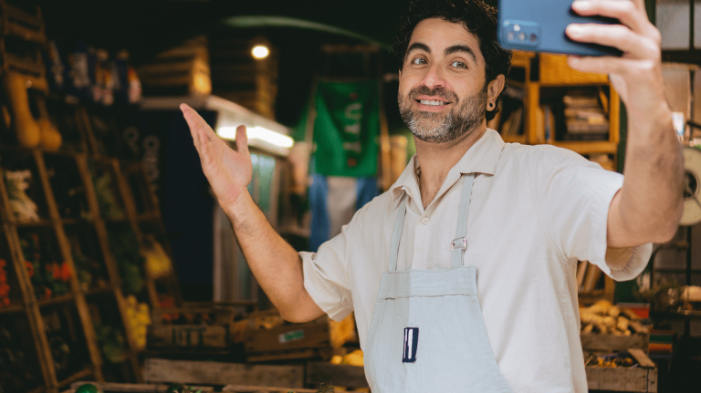 Smiling small business owner wears an apron and takes a selfie in vibrant produce market, amid crates of fresh fruits and vegetables.
