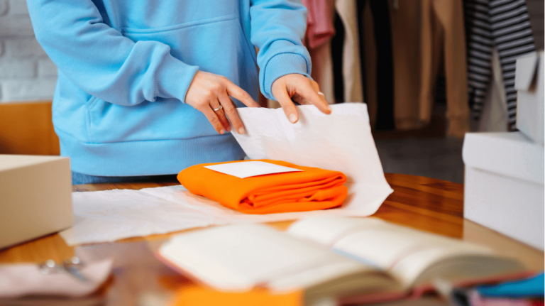 Person in blue hoodie carefully wraps orange clothing in tissue paper at fashion workspace, near hanging garments.