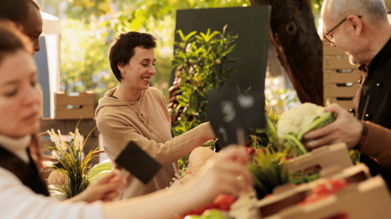Smiling person in beige hoodie reaches for fresh produce at outdoor market, surrounded by vegetables and shaded by sunlit trees.