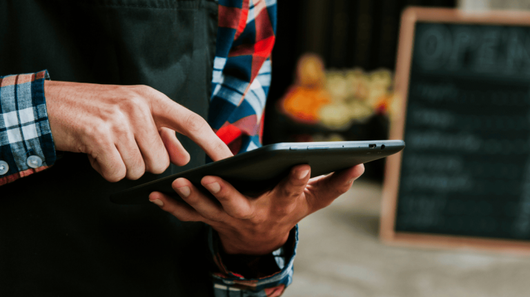 Small business owner in plaid shirt and apron uses tablet at produce stand, managing tasks digitally amid fresh goods and chalkboard signage.