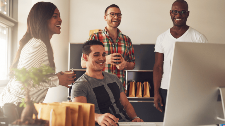 Four casually dressed people gather around a computer in a bright workspace, smiling and collaborating in a relaxed, productive setting.
